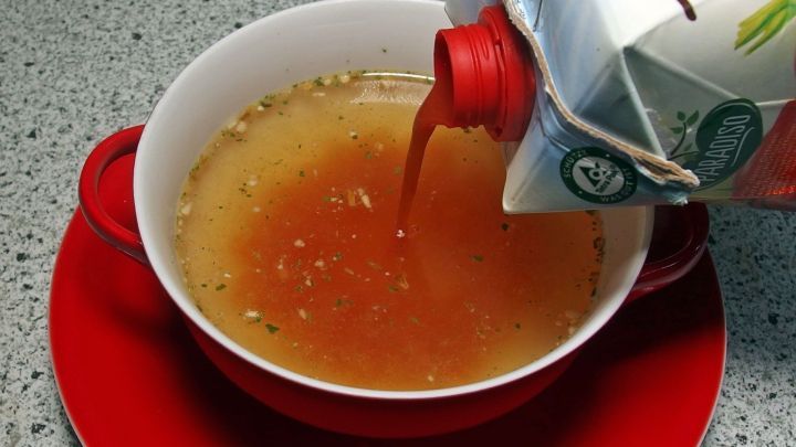 Eine robuste Sch&uuml;sseln mit Tomatensuppe wird gerade aus einem Karton mit Tomatensaft gef&uuml;llt, auf einem grauen Untergrund.