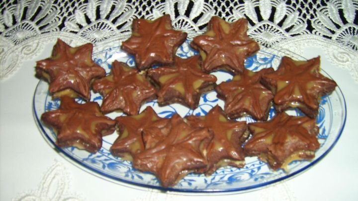 Auf einem Teller liegen schokoladig-gl&auml;nzende Schoko-Karamell-Pl&auml;tzchen in Form von Sternen, umgeben von einem wei&szlig;en Tischtuch.