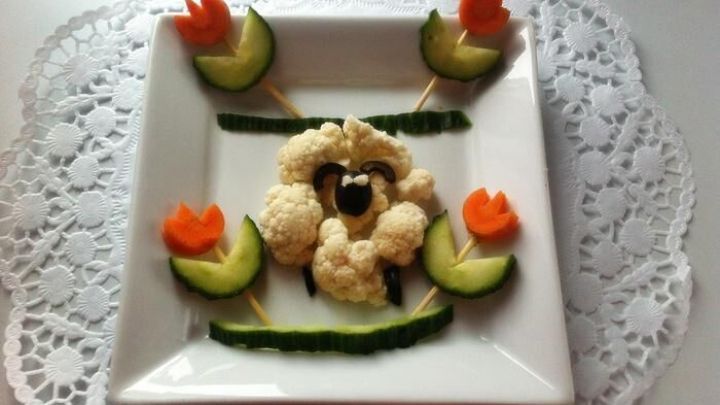 Ein Blumenkohlschaf ist auf einem Teller dekoriert, umgeben von Gurken und Karottenbl&uuml;ten, sch&ouml;n angerichtet.