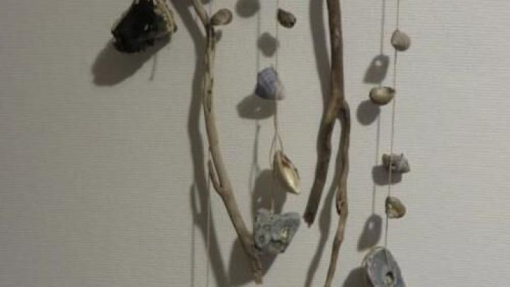 Ein h&ouml;lzerner Mobile h&auml;ngt an einer Wand und zeigt verschiedene Muscheln und Steine, die kunstvoll angeordnet sind.