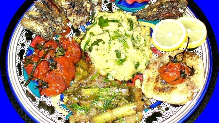 Auf einem dekorativen Teller liegen gebratene Sardinen, Polenta und Backgem&uuml;se, umgeben von frischen Zitronenscheiben.