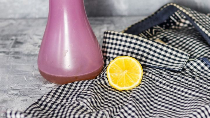 Eine Spr&uuml;hflasche mit pinkem Inhalt steht neben einer halbierten Zitrone und einem karierten Tuch, bereit f&uuml;r die Fleckenentfernung.