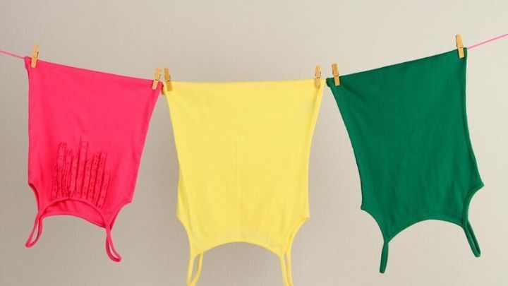 Drei bunte Tanktops (pink, gelb, gr&uuml;n) h&auml;ngen nebeneinander auf einer W&auml;scheleine vor einer neutralen Wand.