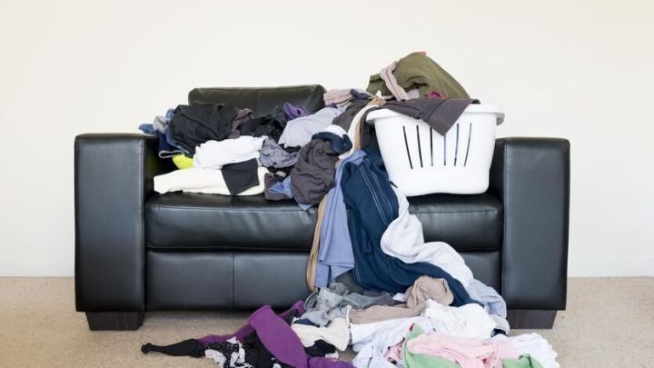 Ein schwarzes Sofa ist mit einem Haufen unordentlicher Kleidung und einem W&auml;schekorb bedeckt, im Wohnzimmer stehend.