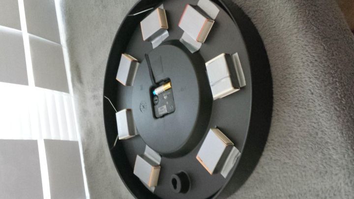 Eine runde, schwarze Uhr mit vielen kleinen, quadratischen Ziffern zeigt die Zeit an. Sie liegt auf einer grauen Oberfl&auml;che.