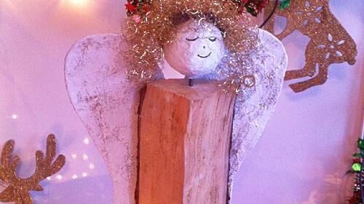 Ein Engel aus Kaminholz steht mit goldener Haarpracht und l&auml;chelt vor einer festlichen, beleuchteten Kulisse.