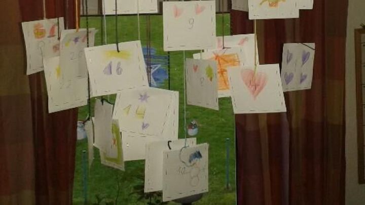 Ein Adventskalender h&auml;ngt an Schn&uuml;ren von einem Holzstab. Die Notizzettel zeigen Zahlen und bunte Zeichnungen vor Vorh&auml;ngen.