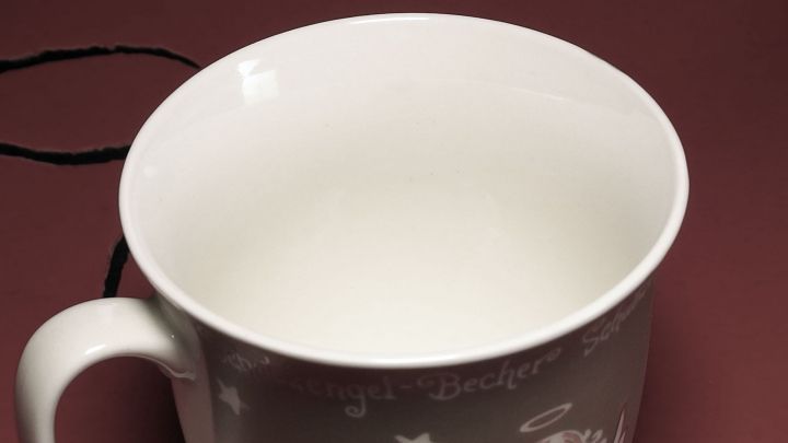 Eine wei&szlig;e Tasse mit Engelsmotiv steht auf einem roten Hintergrund. Rundum ist der Schriftzug "Engel-Becher" in Wei&szlig; zu sehen.
