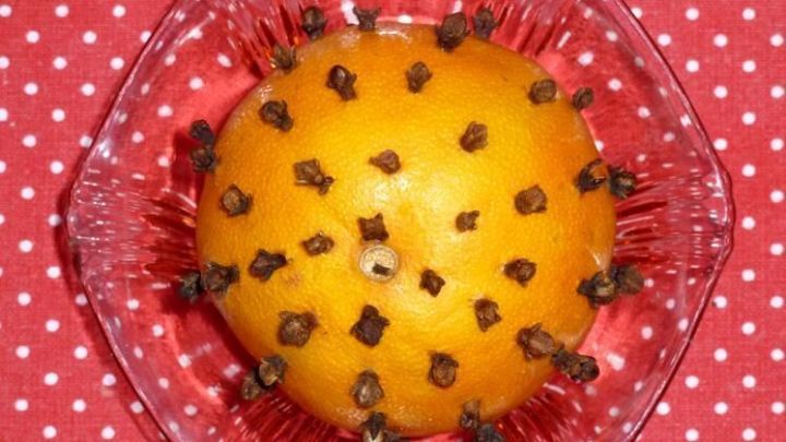 Eine Orange ist mit Nelken best&uuml;ckt und steht in einer klaren Glasschale, umgeben von einem roten gepunkteten Hintergrund.