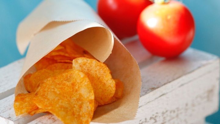 Knusprige Kartoffel-Chips liegen in einer braunen T&uuml;te, neben frischen roten Tomaten auf einem hellen Holzbrett.