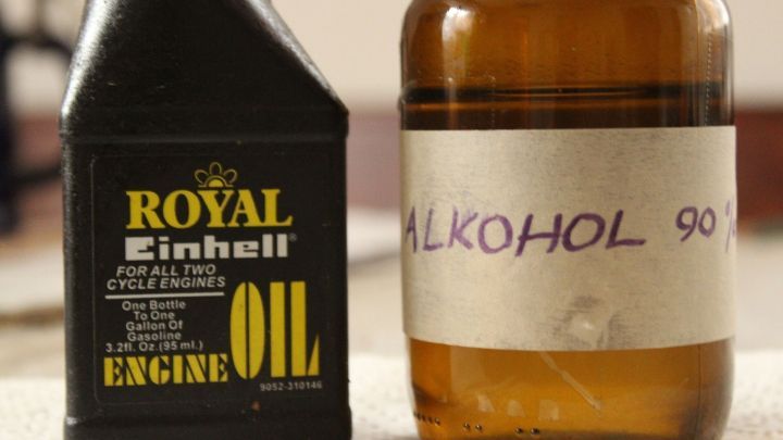 Eine schwarze Flasche mit Maschinen&ouml;l steht neben einer braunen Flasche mit der Aufschrift "ALKOHOL 90&deg;", beide auf einem Tisch.