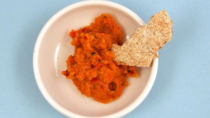 Ein St&uuml;ck Kn&auml;ckebrot dippt in eine orangefarbene, dicke Dipsauce in einer wei&szlig;en Sch&uuml;ssel auf blauem Hintergrund.