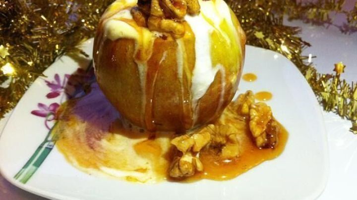 Ein Apfel, gef&uuml;llt mit Eis und karamellisierten Waln&uuml;ssen, steht auf einem Teller und wird von festlichem Goldschmuck umgeben.