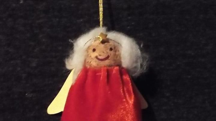 Ein Engel aus Sektkorken h&auml;ngt an einem goldenen Faden, l&auml;chelnd mit rot-goldenen Kleid und wei&szlig;en Haaren vor dunklem Hintergrund.