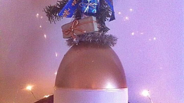 Eine goldene Deko-Weihnachtsglocke h&auml;ngt dekoriert mit Tannenzweigen und Glitzer, umgeben von Lichtern und festlicher Stimmung.