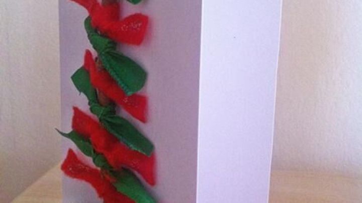 Eine Weihnachtskarte zeigt einen dekorativen Christbaum aus buntem Papier und einem Baumstamm aus Holz, auf einem hellen Tisch.