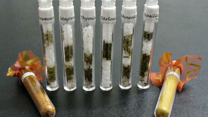 In durchsichtigen Gl&auml;sern pr&auml;sentieren sich Kr&auml;utersalze, die dekorativ mit bunten Schleifen versehen sind, auf einem dunklen Tisch.