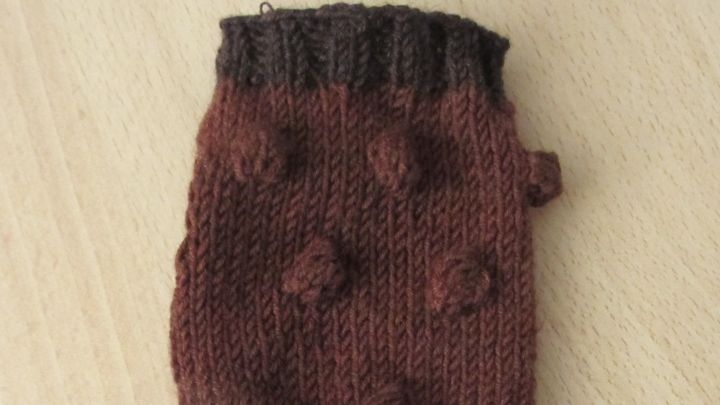 Ein gestrickter Handschuh mit strukturierten Mustern in Braun liegt auf einem hellen Holzuntergrund.