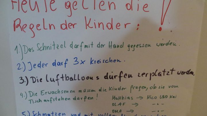 Regeln f&uuml;r Kinder werden an einem Familienessen erkl&auml;rt. Auf einem Plakat stehen f&uuml;nf klare Vorschriften f&uuml;r den Abend. 

Text auf dem Plakat:
"Heute gelten die Regeln der Kinder:
1) Das Schitzel darf mit der Hand gegessen werden.
2) Jeder darf 3x nachholen.
3) Die Luftballons d&uuml;rfen zerplatzt werden.
4) Die Erwachsenen m&uuml;ssen die Kinder fragen, ob sie noch Hunger haben.
5) Schokolade wird, wenn genug da ist, verteilt."