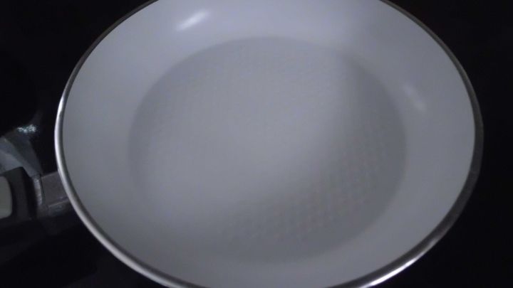 Eine wei&szlig;e Keramikpfanne liegt auf einem schwarzen Kochfeld, bereit zum Kochen oder Braten.