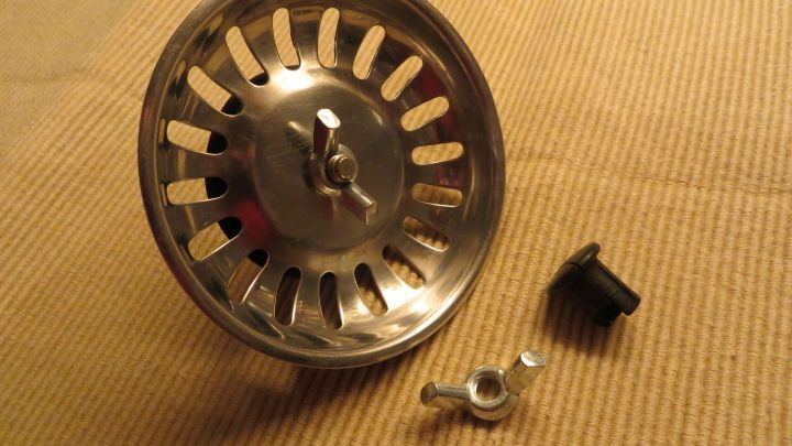 Ein Siebkorb liegt auf einer abatoirf&ouml;rmigen Oberfl&auml;che, w&auml;hrend ein kaputter Plastikschraubkopf daneben liegt, bereit zum Austausch.