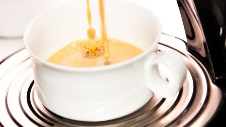 Eine Tasse wird mit frisch gebr&uuml;htem Kaffee gef&uuml;llt, w&auml;hrend sie auf einer Ablage neben einer Kaffeemaschine steht.