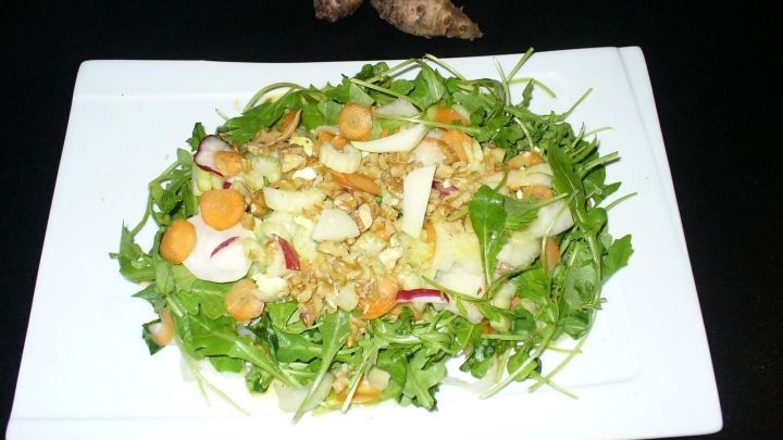 Ein frischer Topinambursalat mit gr&uuml;nen Bl&auml;ttern, N&uuml;ssen und gehobeltem Gem&uuml;se auf einem wei&szlig;en Teller, umgeben von dunklem Hintergrund.