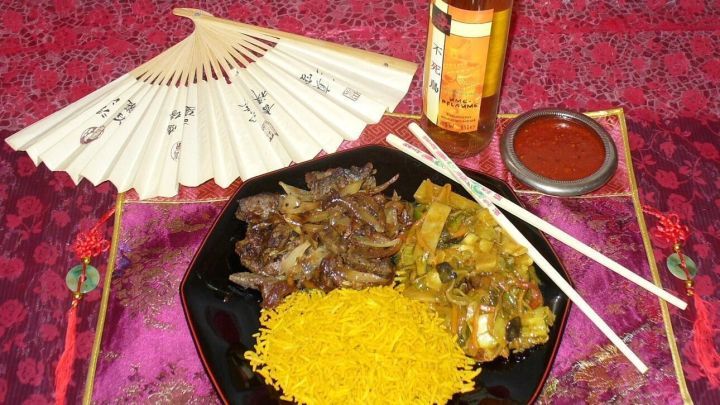 Rindfleisch liegt auf einem schwarzen Teller, umgeben von asiatischem Gem&uuml;se und gelbem Reis, mit St&auml;bchen und einem Getr&auml;nk.