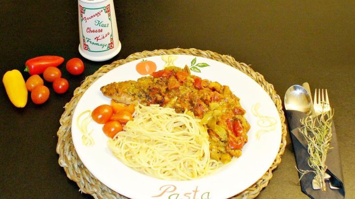 Ein &uuml;berbackenes Pesto-Putenschnitzel liegt auf einem Teller mit Pasta, umgeben von frischen Tomaten und einer Gew&uuml;rzdose.