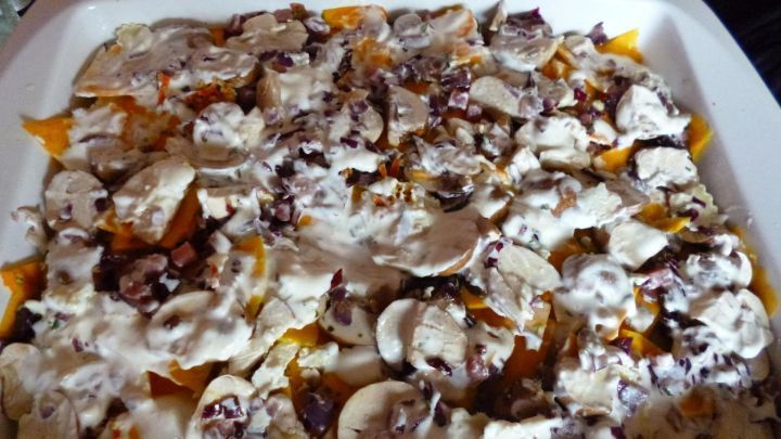 Eine Auflaufform, gef&uuml;llt mit K&uuml;rbislasagne, ist mit Schicht von Sahne und Pilzen bedeckt. Sie steht in einer K&uuml;che.