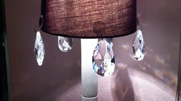 Eine alte Tischlampe steht auf einem wei&szlig;en Tisch und wird mit einem dunkelbraunen Lampenschirm und Glaskristallen aufgepeppt.