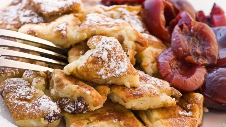 Zarte St&uuml;cke von Grie&szlig;schmarrn liegen auf einem Teller, umgeben von fruchtigem Kompott. Ein Besteck greift nach dem Essen.