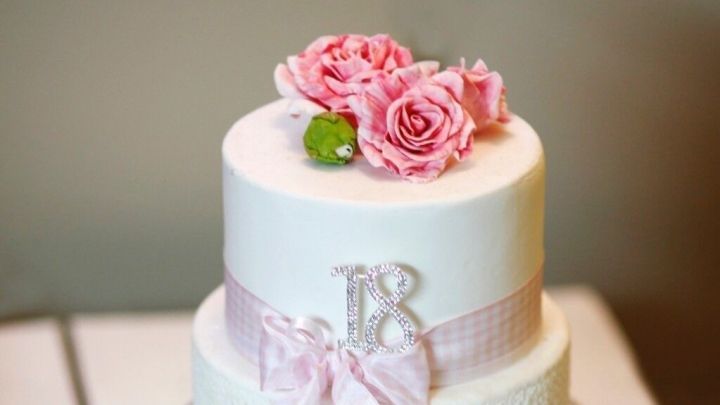 Ein dekorierter Kuchen aus Rollfondant steht auf einem wei&szlig;en Tortenteller, umgeben von rosa Blumen und einem zarten Band.