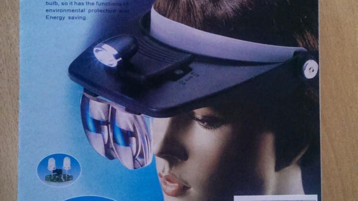 Eine Stirnlupe mit Licht liegt auf einem Tisch, w&auml;hrend sie auf einem Modellkopf abgebildet ist, ideal f&uuml;r feine handwerkliche Arbeiten.