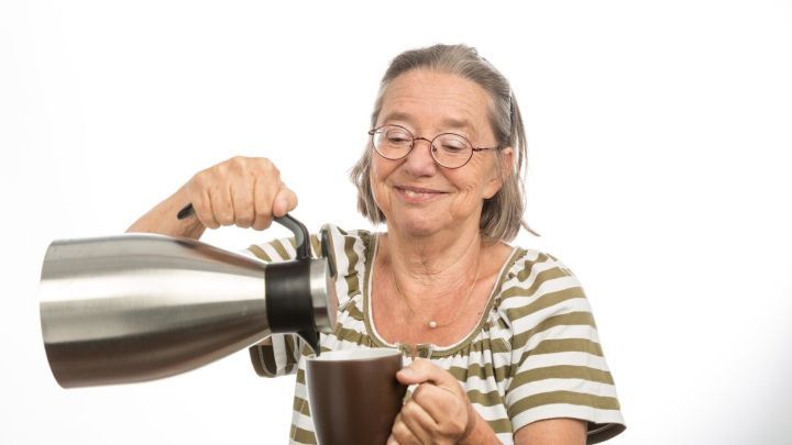 Eine &auml;ltere Dame gie&szlig;t mit einem Edelstahl-Thermoskanne l&auml;chelnd eine warme Fl&uuml;ssigkeit in eine Tasse, vor neutralem Hintergrund.