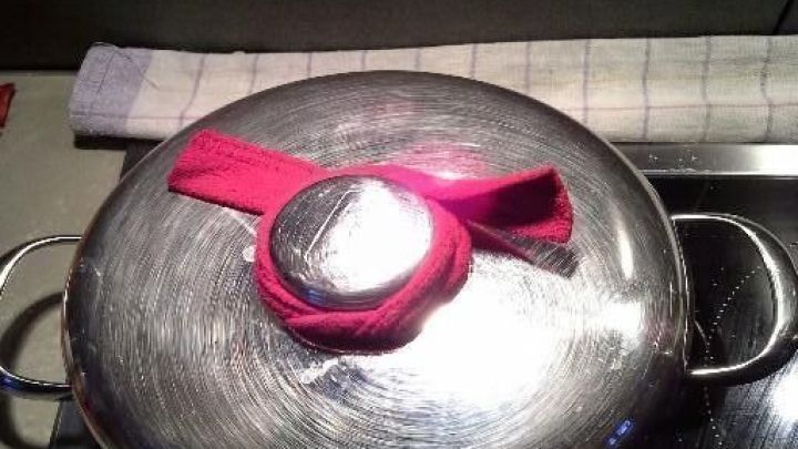 Ein hei&szlig;er Kochtopfdeckel liegt auf einem Herd, mit einem pinken Tuch als Schutz vor der Hitze im K&uuml;chenumfeld.