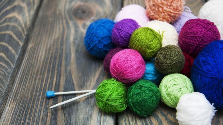 Bunte Wollkn&auml;uel liegen auf einem holzernen Tisch, w&auml;hrend Stricknadeln darauf warten, verwendet zu werden.