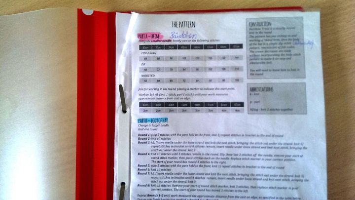 Eine rote Mappe liegt auf einem Tisch, in der Dokumente sortiert sind, die Anleitungen f&uuml;r das Stricken enthalten.