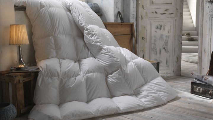 Ein gro&szlig;es, wei&szlig;es Daunenbett liegt schr&auml;g an einer Wand in einem lichtdurchfluteten Raum mit Holzboden und einer gem&uuml;tlichen Lampe.