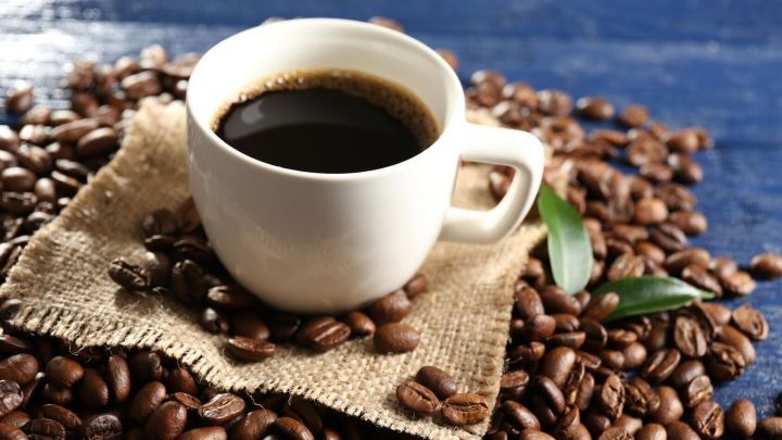 Eine dampfende Tasse Kaffee steht auf einem Jutesack, umgeben von gro&szlig;z&uuml;gigen Kaffeebohnen auf einem blauen Hintergrund.