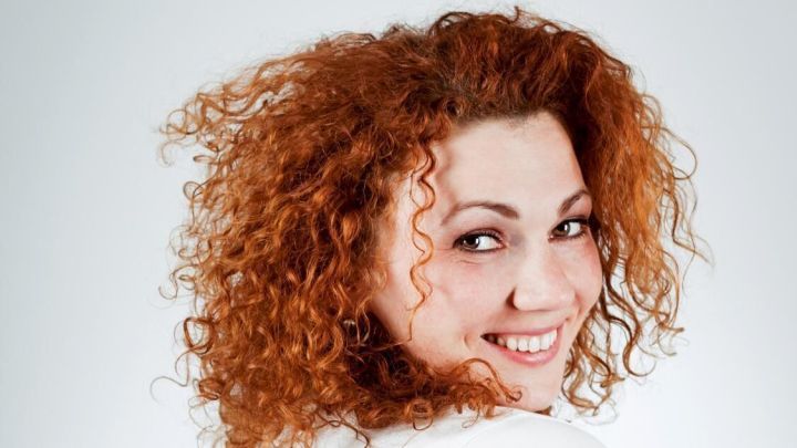 Eine lachende Frau mit lockigem, krausem Haar blickt &uuml;ber die Schulter in die Kamera, vor einem neutralen Hintergrund.