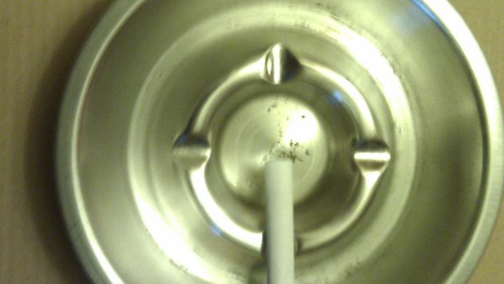 Ein runder Aschenbecher h&auml;ngt an der Wand, w&auml;hrend eine Zigarette sicher darin positioniert ist, um ein Herunterfallen zu verhindern.