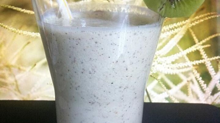 Ein cremiger Kiwi-Shake mit Honig steht in einem Glas, umgeben von einem hellen, pflanzlichen Hintergrund.