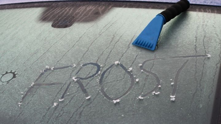 Eine Eisschicht auf einer Autoscheibe wird mit einem blauen Eiskratzer bearbeitet, w&auml;hrend das Wort &bdquo;FROST&ldquo; sichtbar ist.