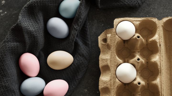 Bunte Ostereier aus Naturmaterialien liegen auf einem dunklen Tuch neben einem Eierkarton, der leere Eier zeigt.