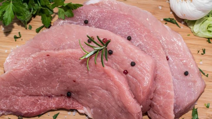 Drei saftige Fleischst&uuml;cke liegen auf einem Holzbrett, umgeben von Kr&auml;utern wie Rosmarin und Knoblauch, bereit f&uuml;r Marinade.