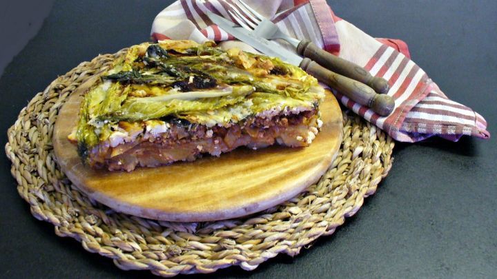 Ein Kohlauflauf mit Fetak&auml;se liegt auf einem Holzbrett, umgeben von einem geflochtenen Untersetzer und einer gestreiften Serviette.