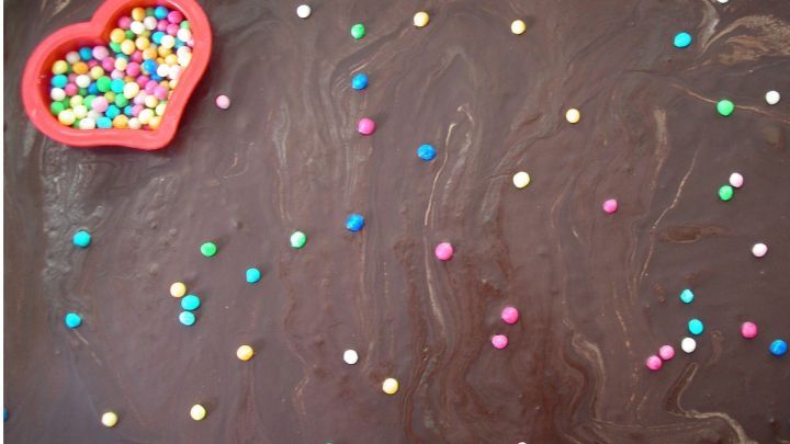 Ein herzf&ouml;rmiger Beh&auml;lter mit bunten Streuseln liegt auf einer Schokoladenschicht, die mit Spa&szlig; dekoriert wird.