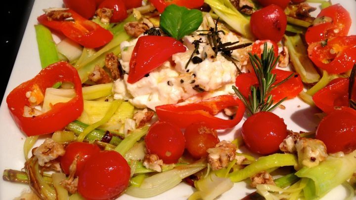 Gebackener Schafsk&auml;se liegt auf einem bunten Bett aus karamellisiertem Lauch, umgeben von roten Paprika und Tomaten.