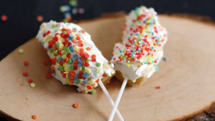 Zwei Bananen am Stiel sind mit Creme und bunten Streuseln dekoriert, auf einem Holzbrett vor dunklem Hintergrund.